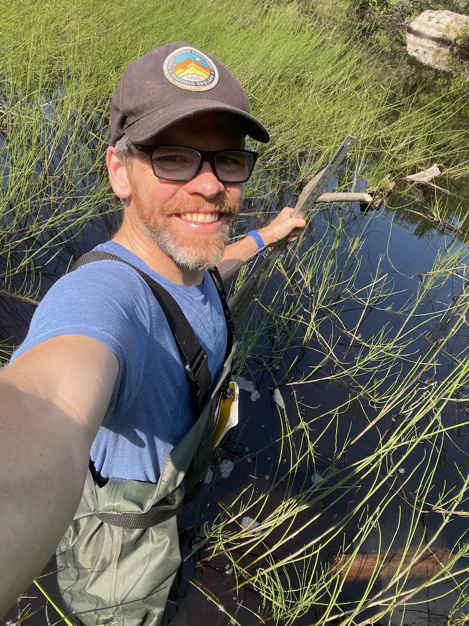 Dr. Adam Algar Doing Fieldwork Dr. Adam Algar stands in marsh wearing hip waders and holding a measuring stick