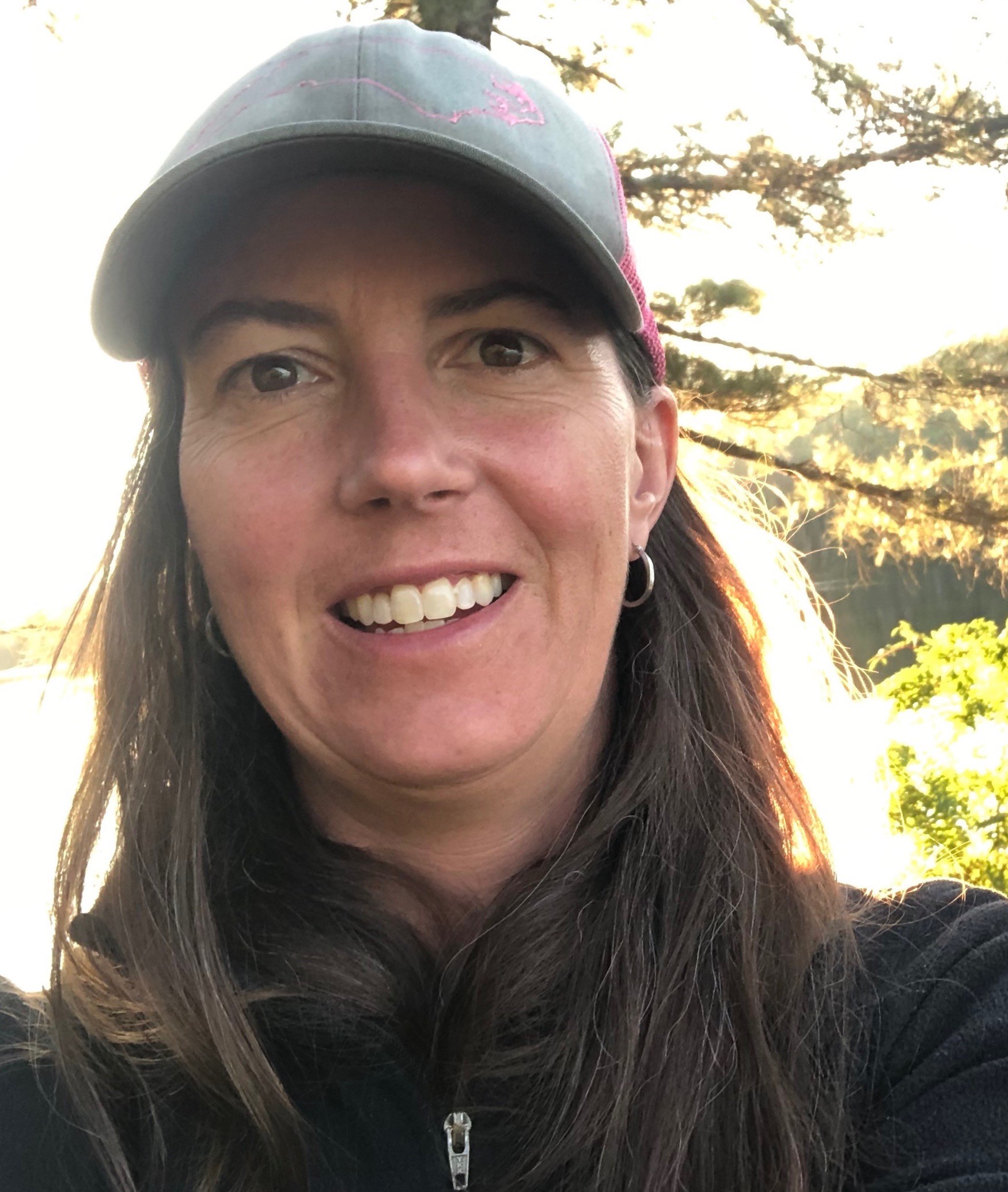 Lindsay Sargent A head-and-shoulders photo of Linsday Sargent outside wearing a grey fleece zip-up, baseball cap, and hoop earring; a tree and a lake are visible in the background