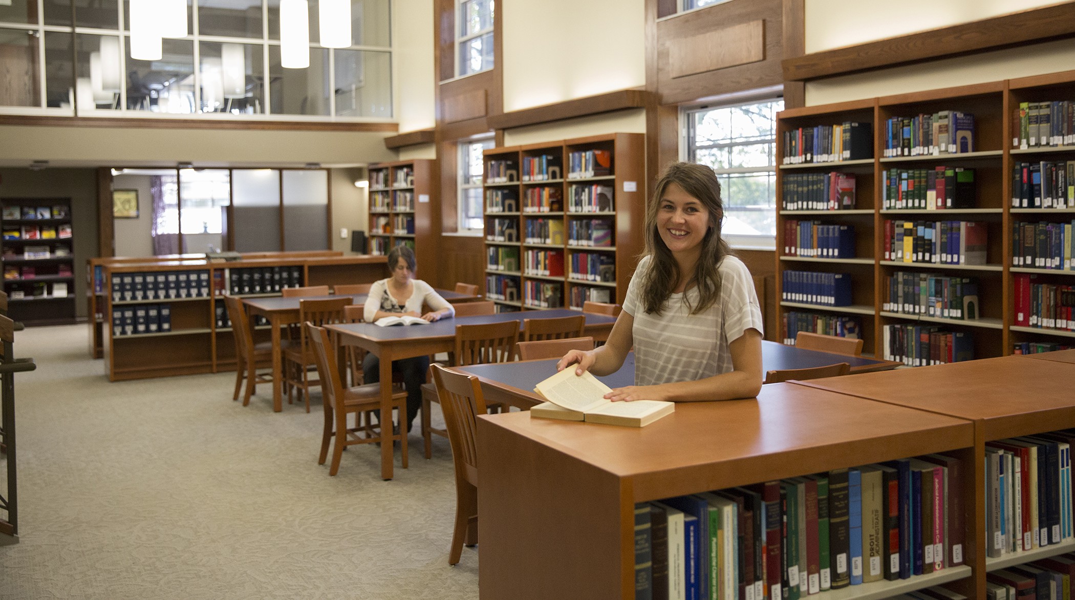 Bora Laskin Law Library An interior view of the Bora Laskin Faculty of Law Library with a young woman standing behind a low bookcase with an open book and a seated woman reading a book at a table in the background