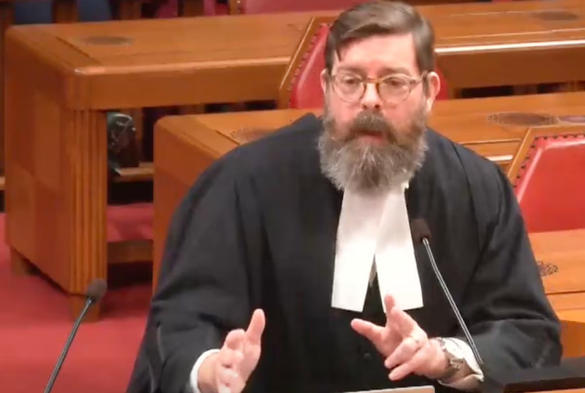Ryan Alford in Supreme Court Ryan Alford, wearing glasses, a black robe, and a white jabot, sits a table with two microphones in a red-carpeted courtroom with empty wooden tables and chairs behind him