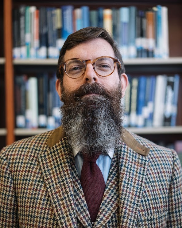 Ryan Alford with Bookcase Ryan Alford, wearing glasses, a tie, and a houndstooth-checked vest and blazer, stands in front of a bookcase