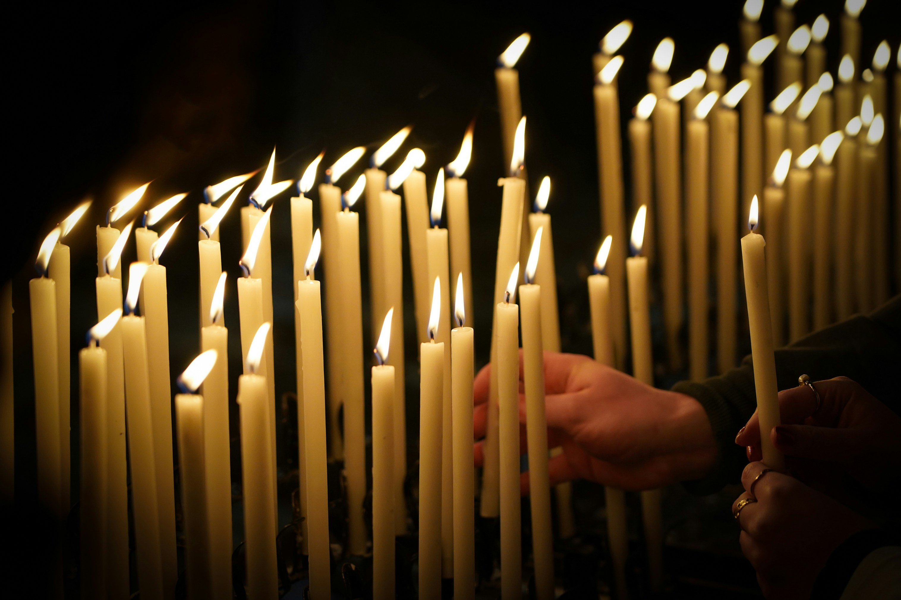 Candles The hands of two people holding lit candles add their candles to a group of white candles burning in the darkness