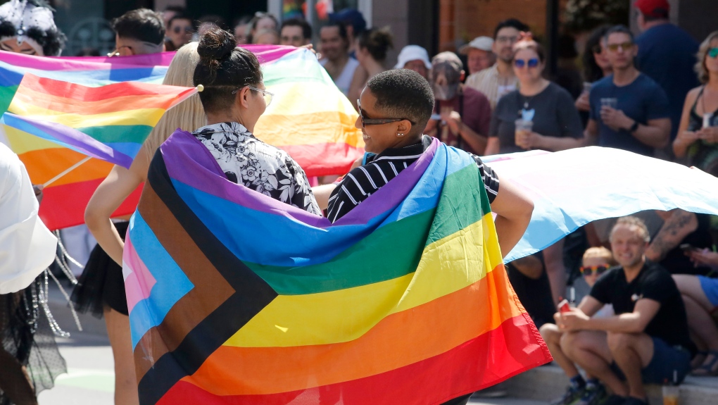 Two individuals wearing a Progress Pride flag draped over their shoulders, stand facing each other at a Pride Parade. 