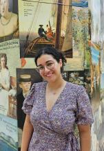 A woman wearing a purple dress, black curly hair tied away from the face and wearing glasses