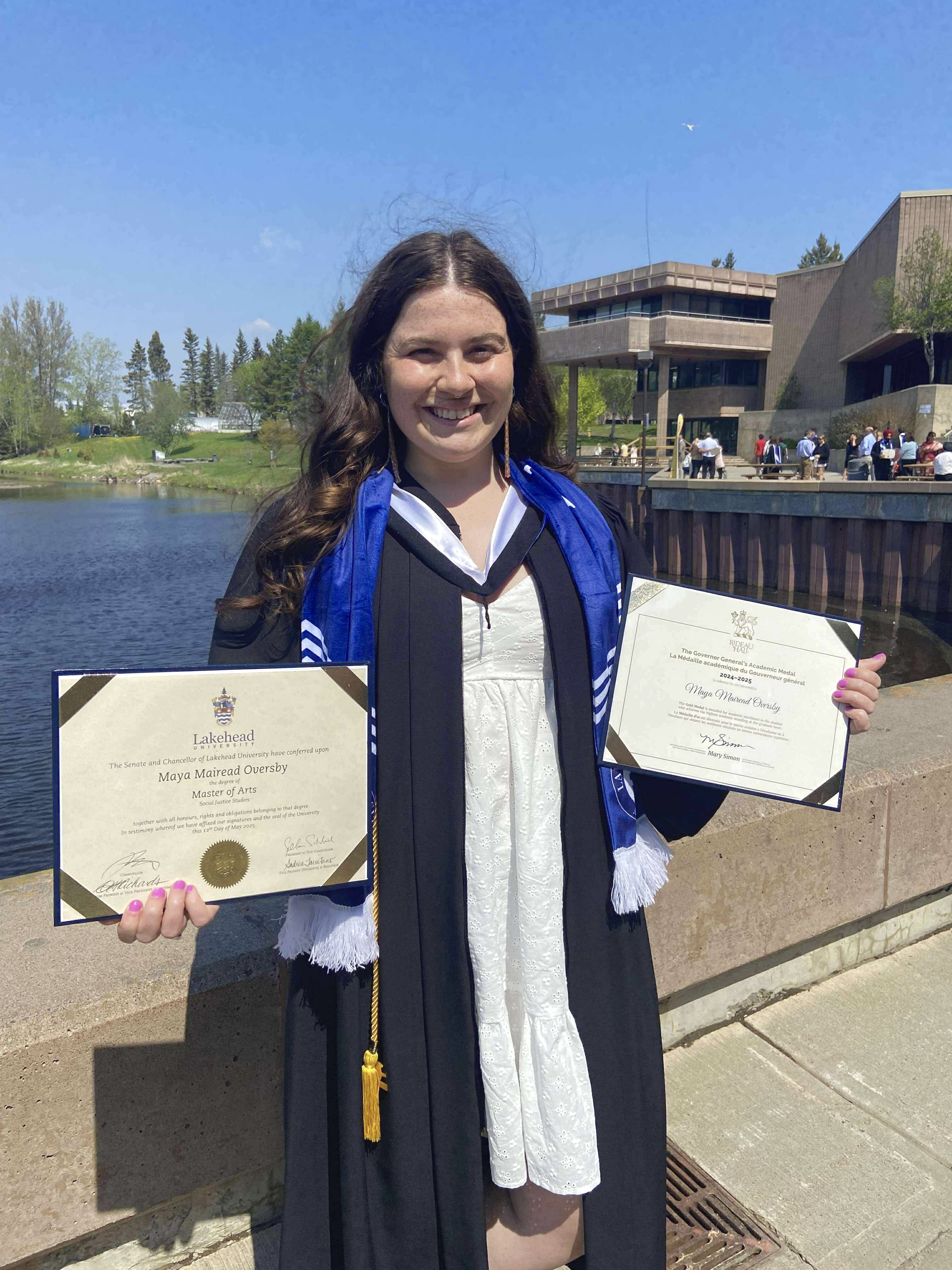 Maya Oversby, Award Winner. Maya Oversby holding two awards in front of Lake Tamblyn, Lakehead University campus.